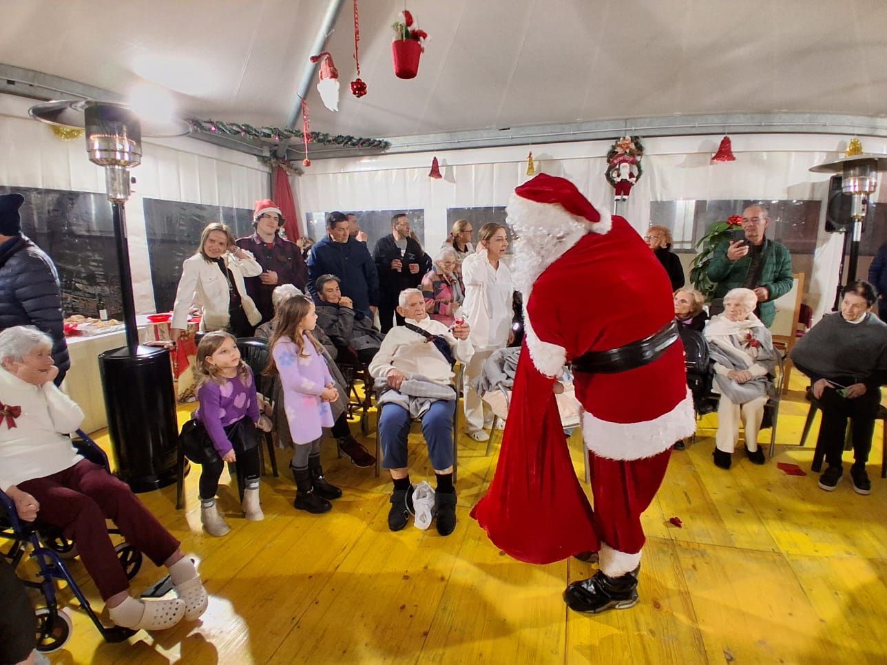 Babbo Natale tiene in mano un sacco rosso in una stanza decorata, mentre un gruppo di persone lo osserva, alcune sedute e altre in piedi.