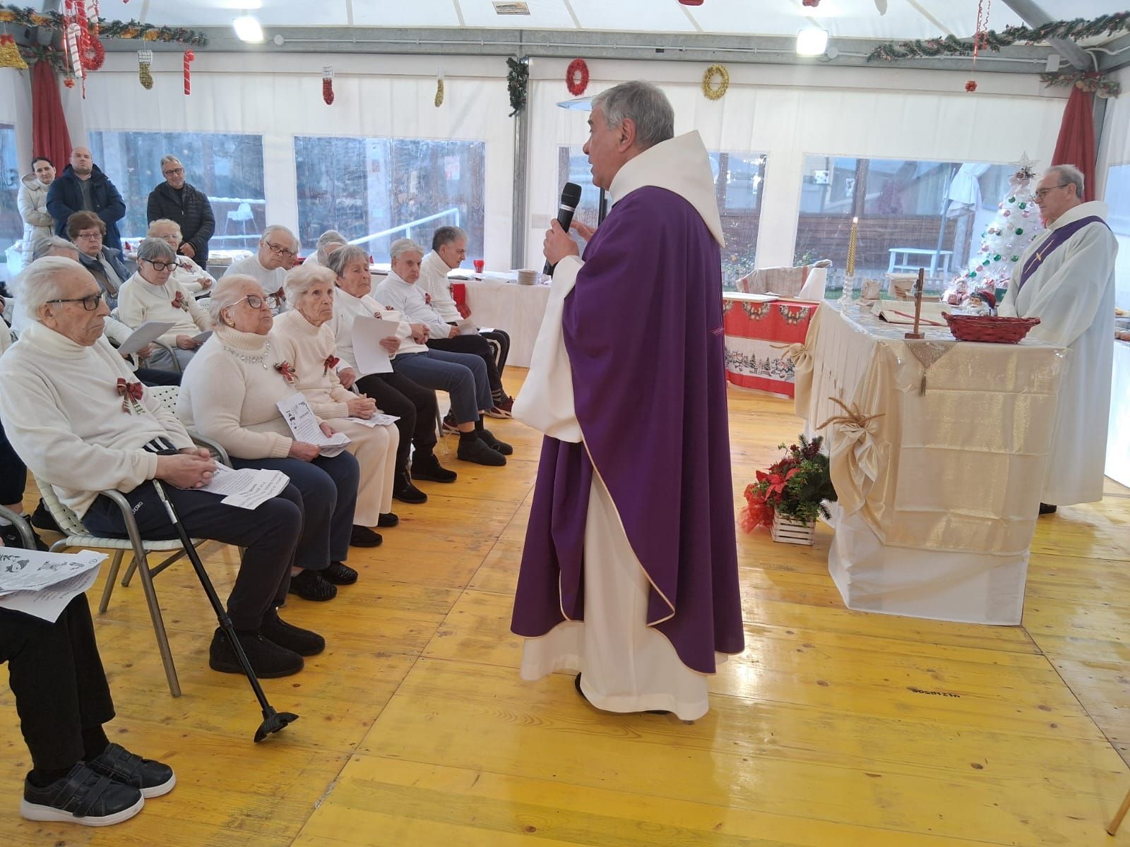 Un sacerdote vestito di viola celebra una funzione religiosa per un pubblico seduto in una sala luminosa simile a una tenda.