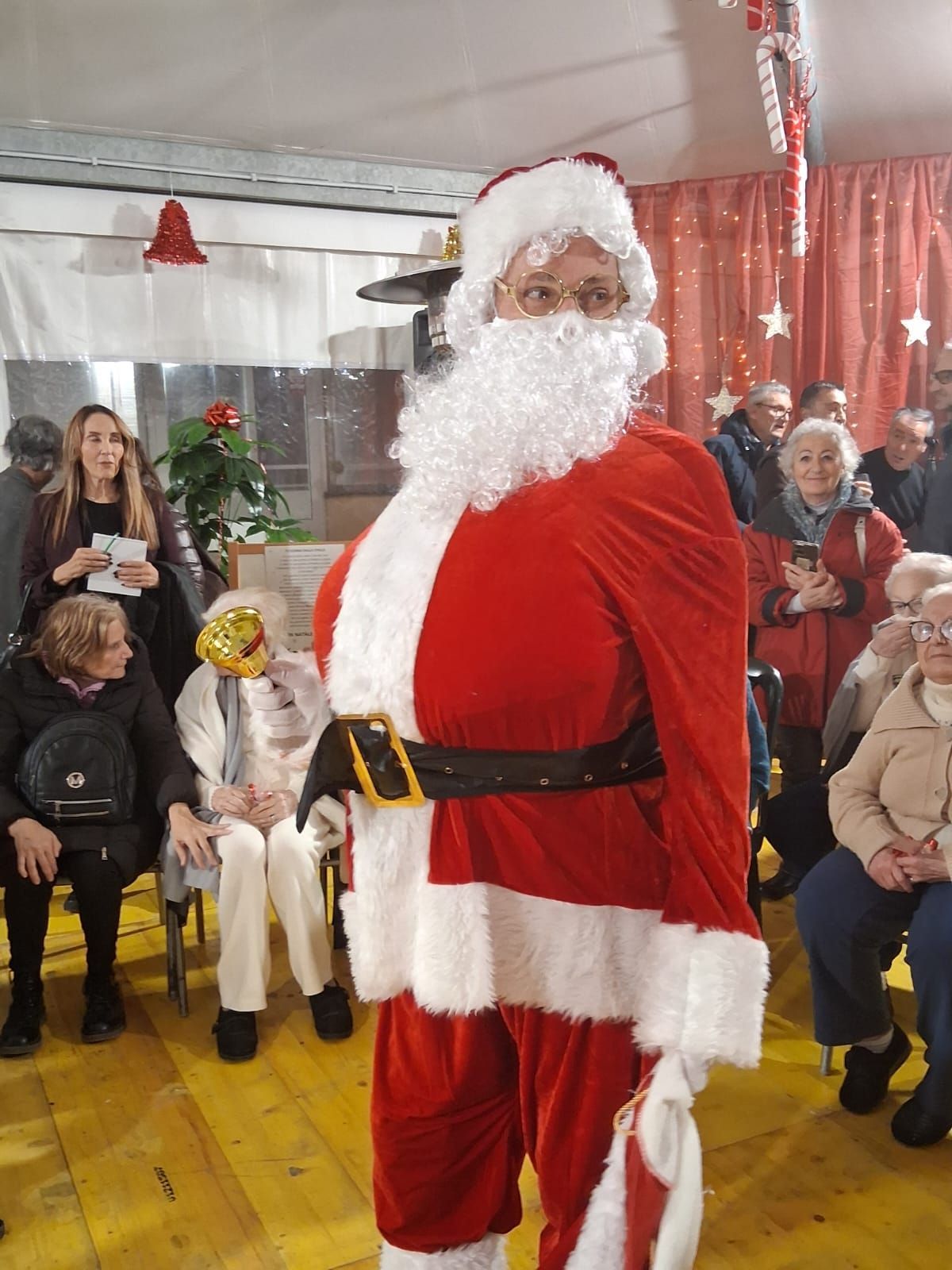 Una persona vestita da Babbo Natale si trova in una stanza decorata, circondata da un pubblico seduto su delle sedie.