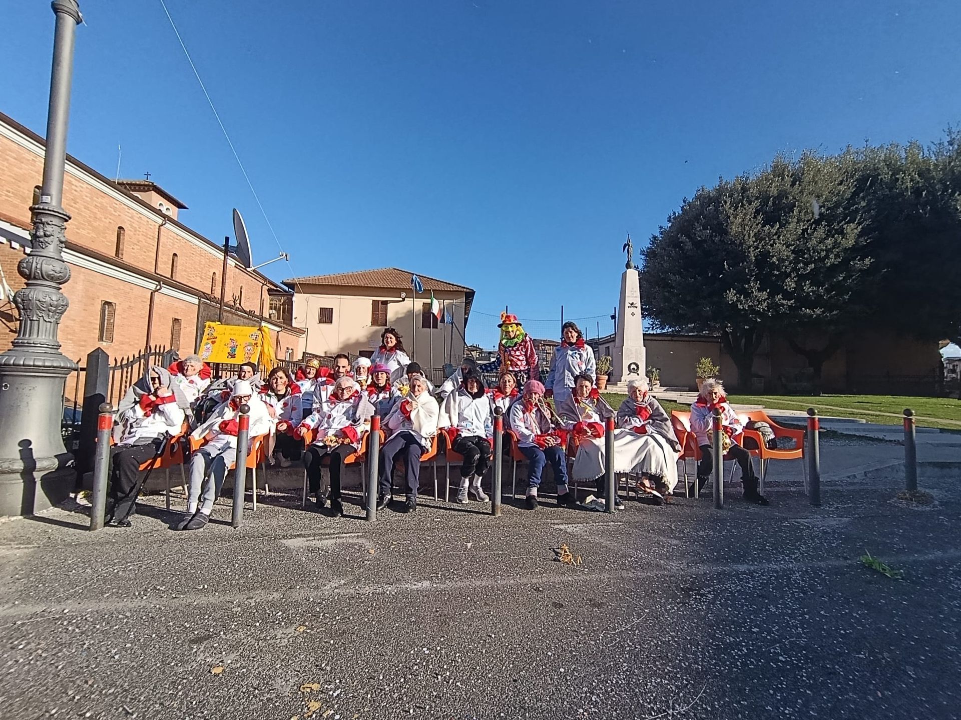 Un gruppo di persone con indosso sciarpe rosse siede sulle panchine in una piazza italiana sotto un cielo azzurro e limpido.