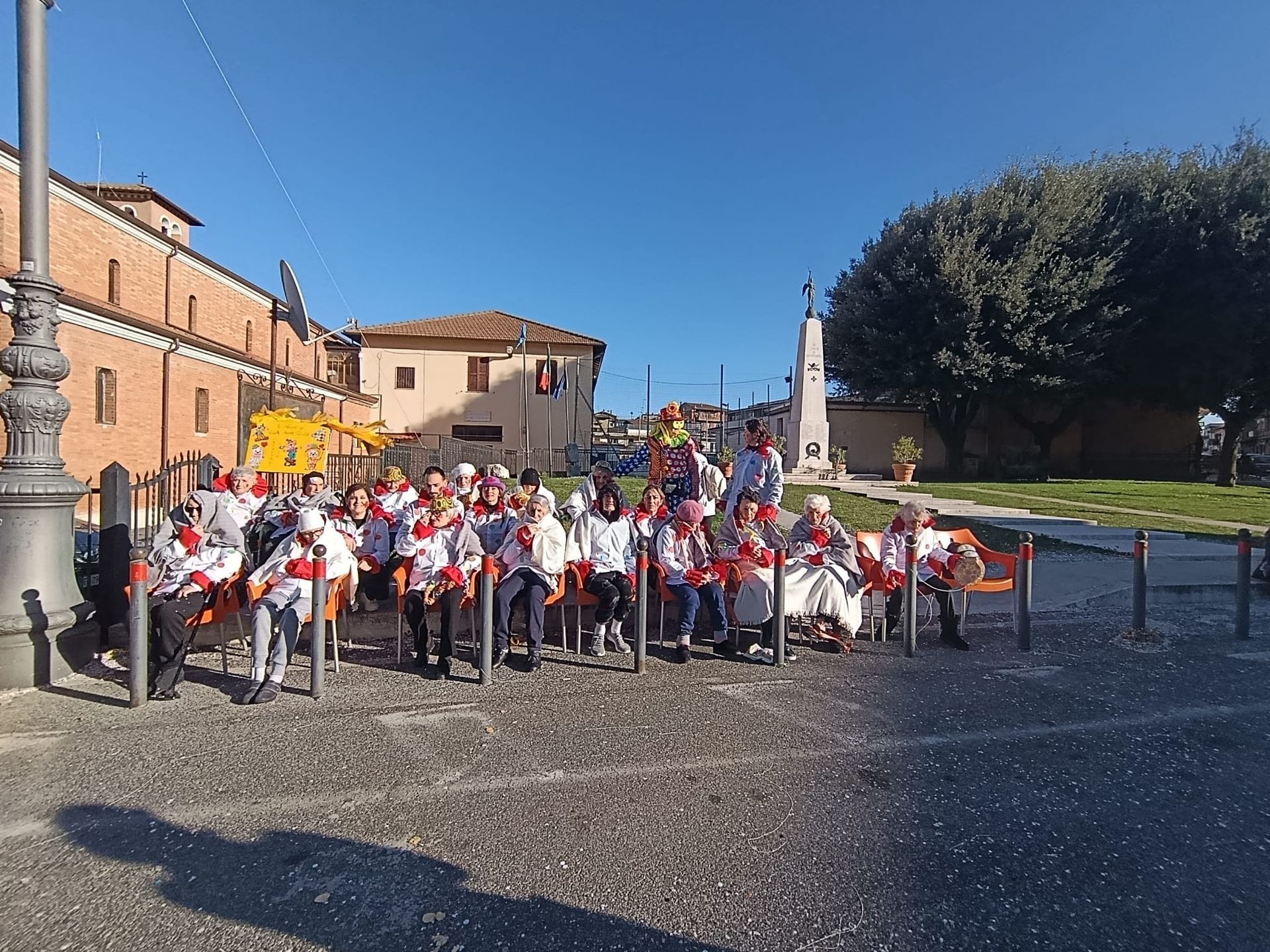 Un gruppo di persone con indosso magliette bianche è seduto su delle sedie all'aperto in una piazza lastricata di fronte a un edificio color beige e a un monumento.