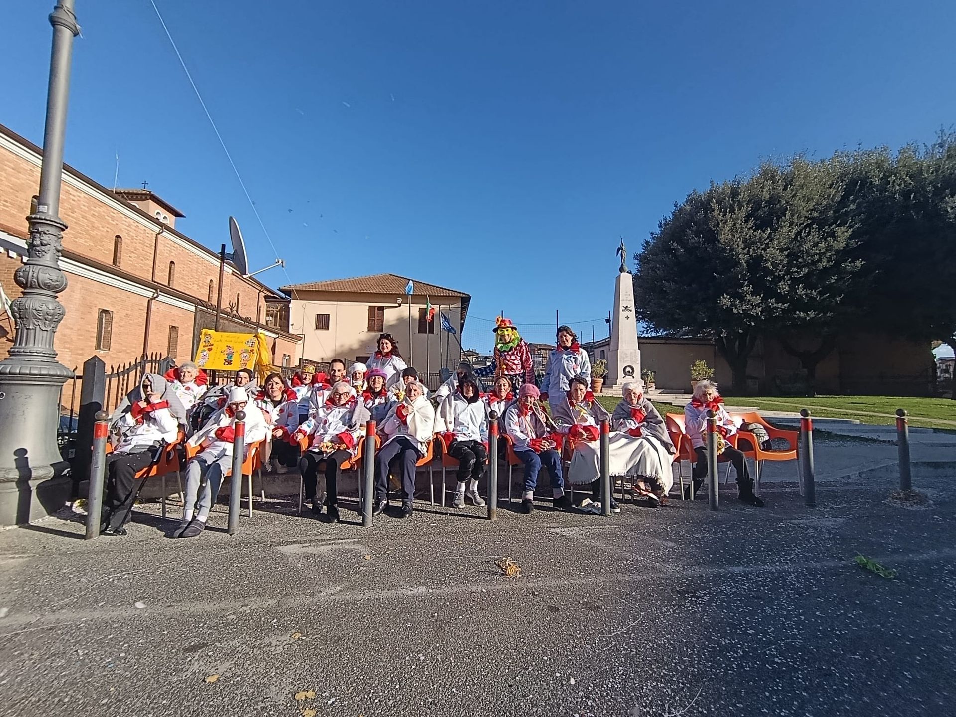 Un folto gruppo di persone vestite di bianco siede su delle sedie in una piazza soleggiata, posando per una foto vicino a un alto monumento.
