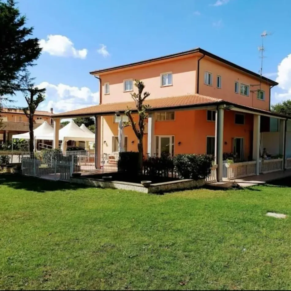 Una casa arancione a due piani con una veranda che la circonda completamente e tende bianche in giardino, sullo sfondo di un cielo azzurro con nuvole bianche.