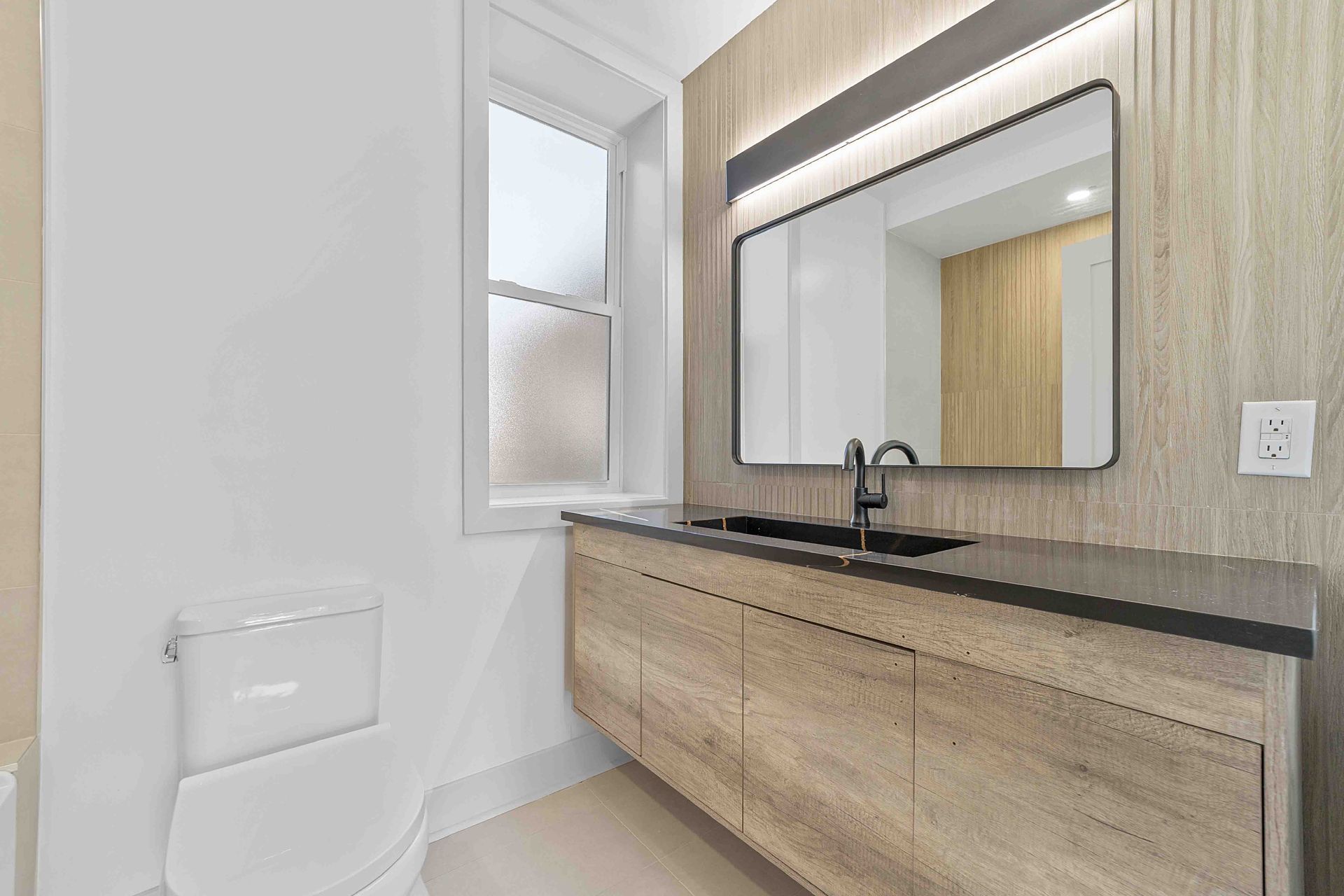 A bathroom with a toilet , sink , mirror and window.