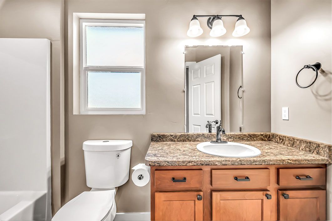 A bathroom with a toilet , sink , mirror and window.