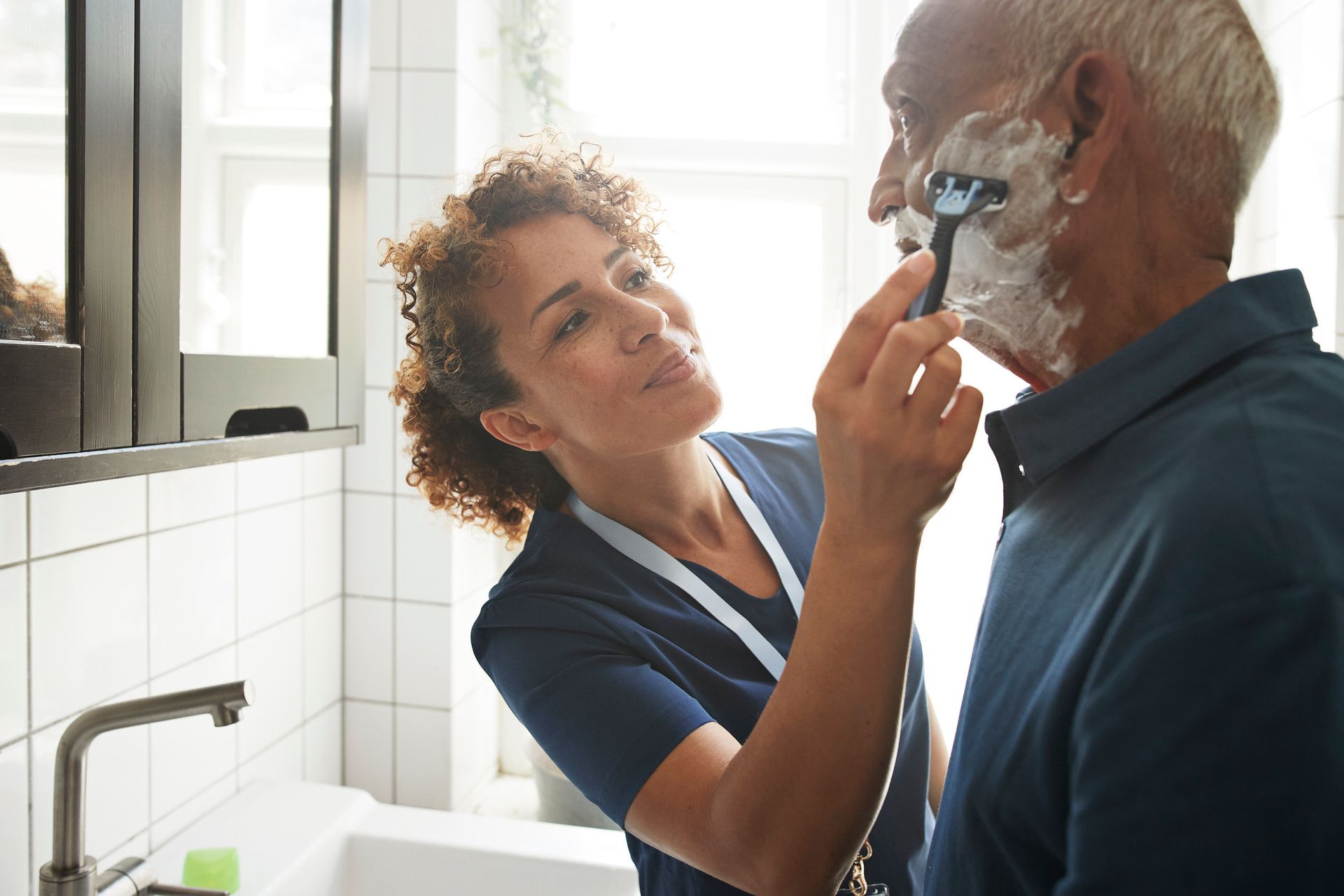 A nurse is shaving an older man 's face in a bathroom.