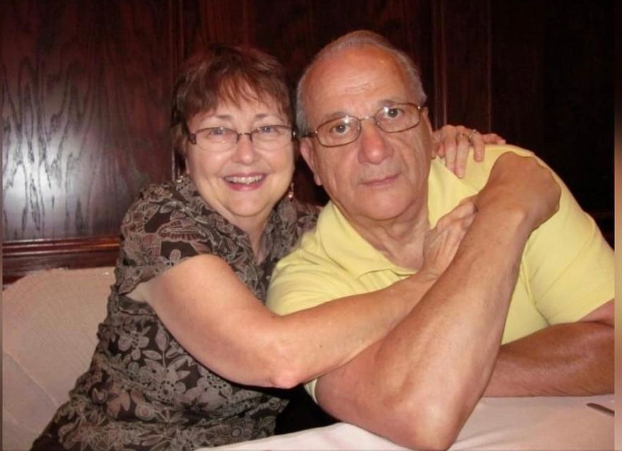 Couple smiling, embracing at a table. Woman in patterned blouse; man in yellow polo. Brown background.