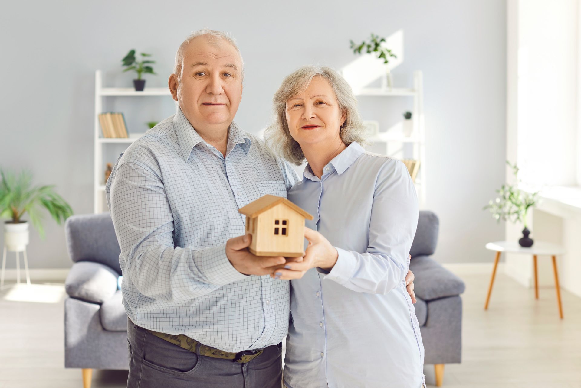 Older couple holding a small wooden house. They smile in a living room with a couch and plants.