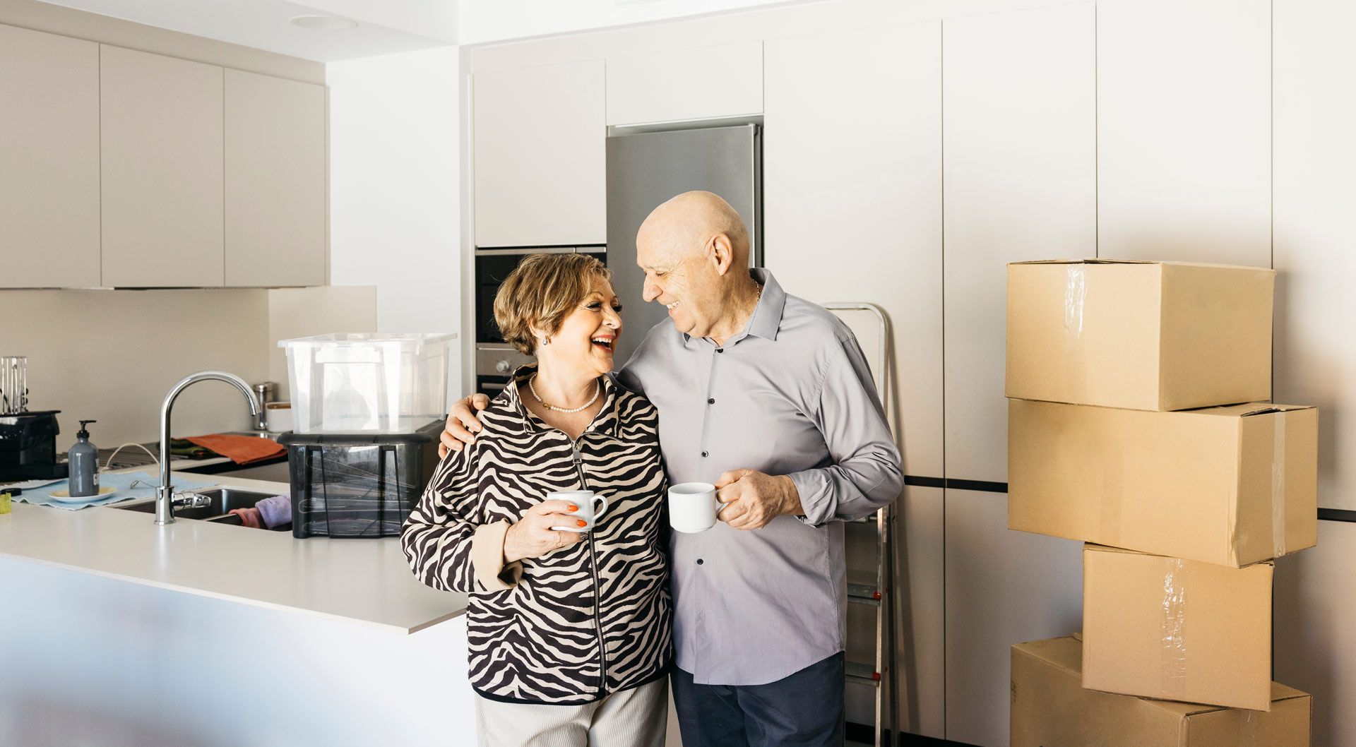 Elderly couple in kitchen, arms around each other, smiling, holding mugs, boxes stacked nearby.