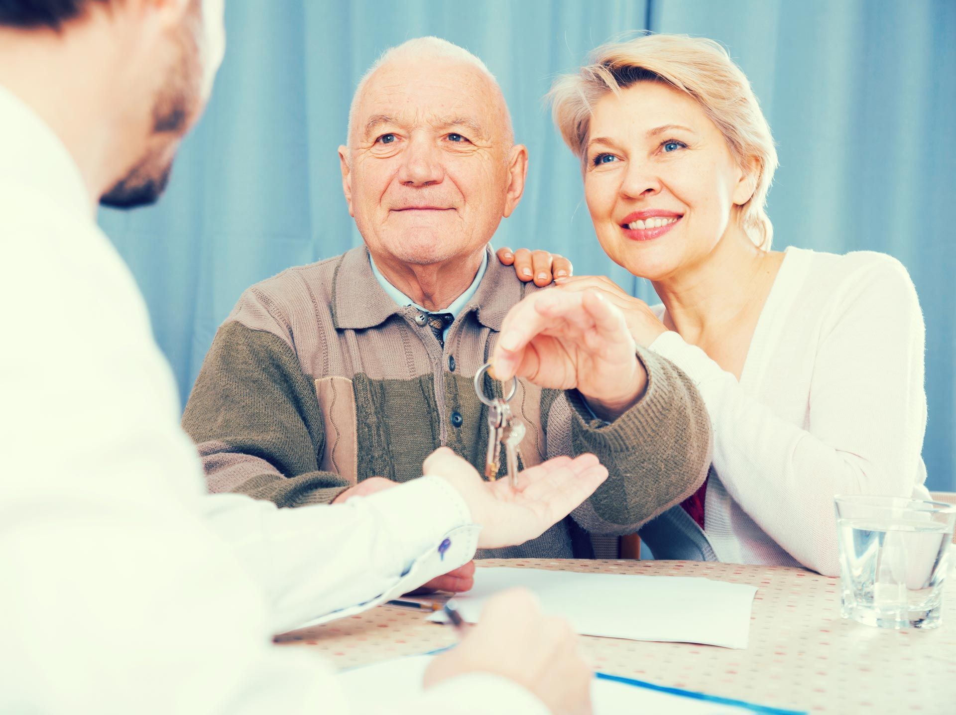 A person handing keys to an older couple seated at a table, smiling.