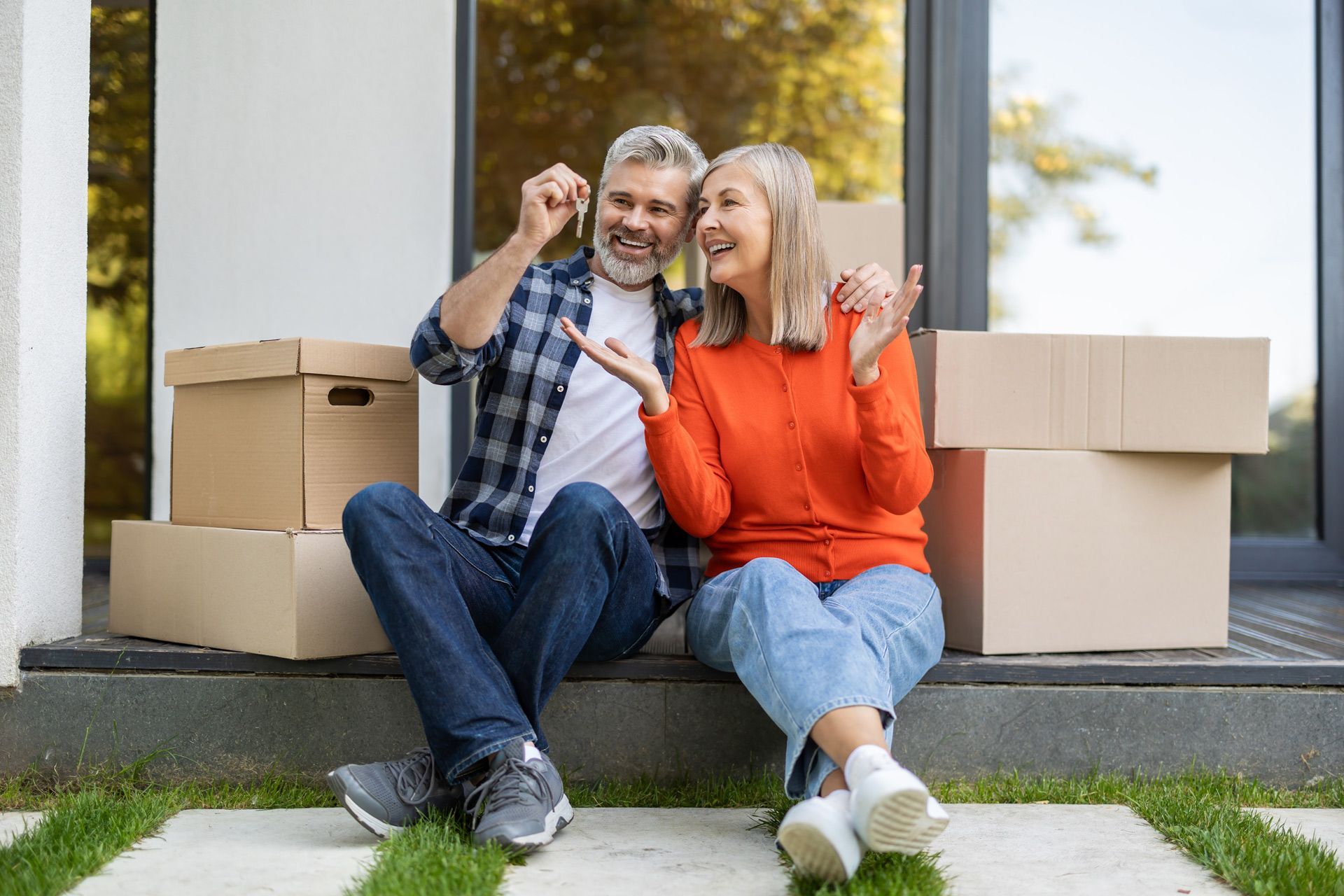 Couple sitting on steps, holding keys, surrounded by moving boxes; happy expressions.