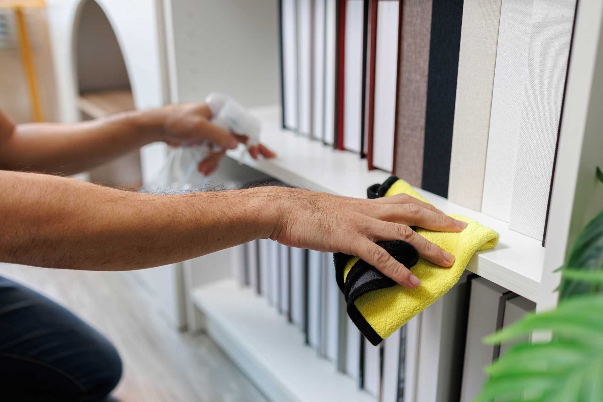 Person cleaning white bookshelf with a spray bottle and yellow cloth.