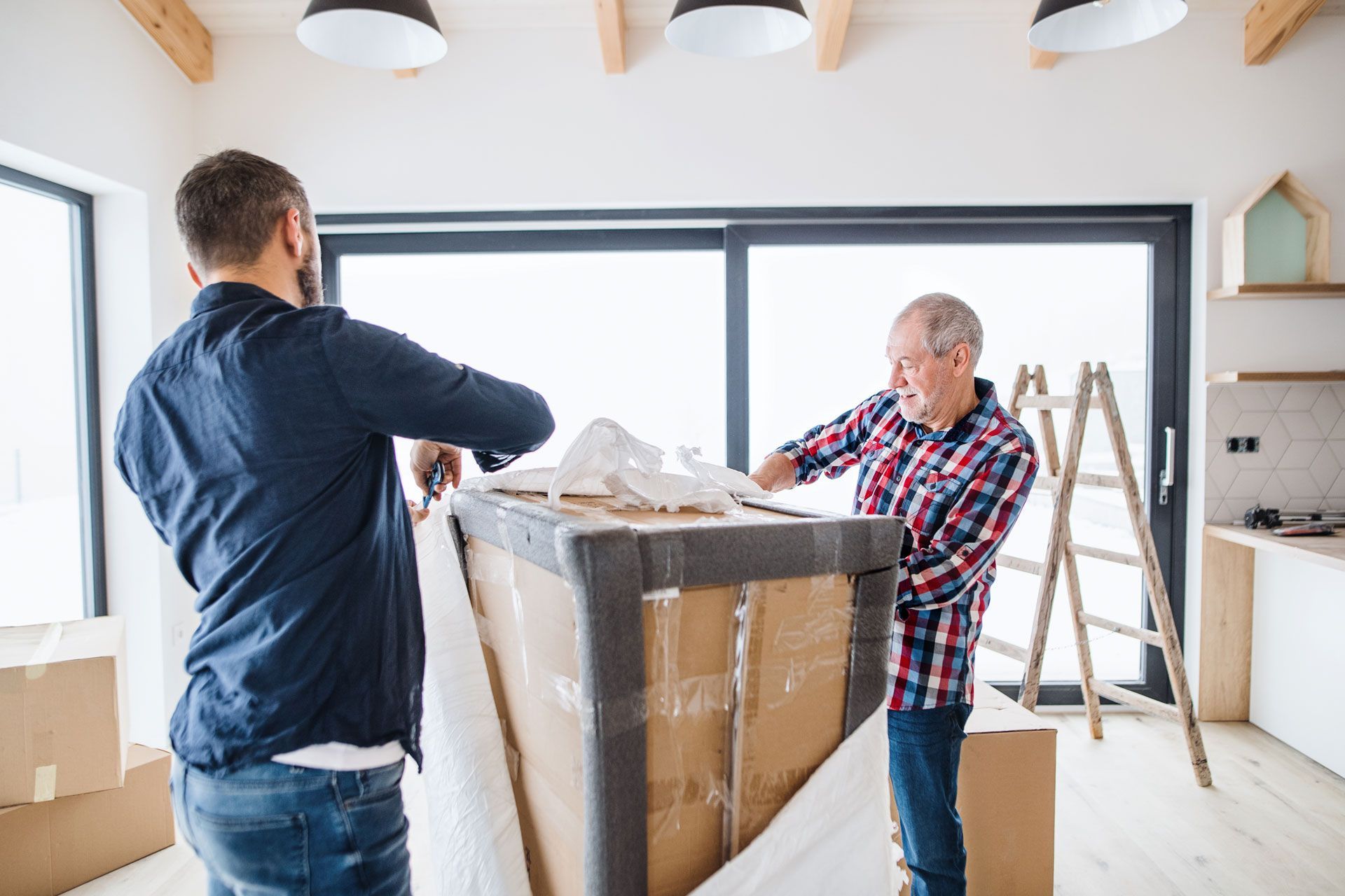 Two people unwrapping a large box in a light-filled room with moving boxes and a ladder visible.