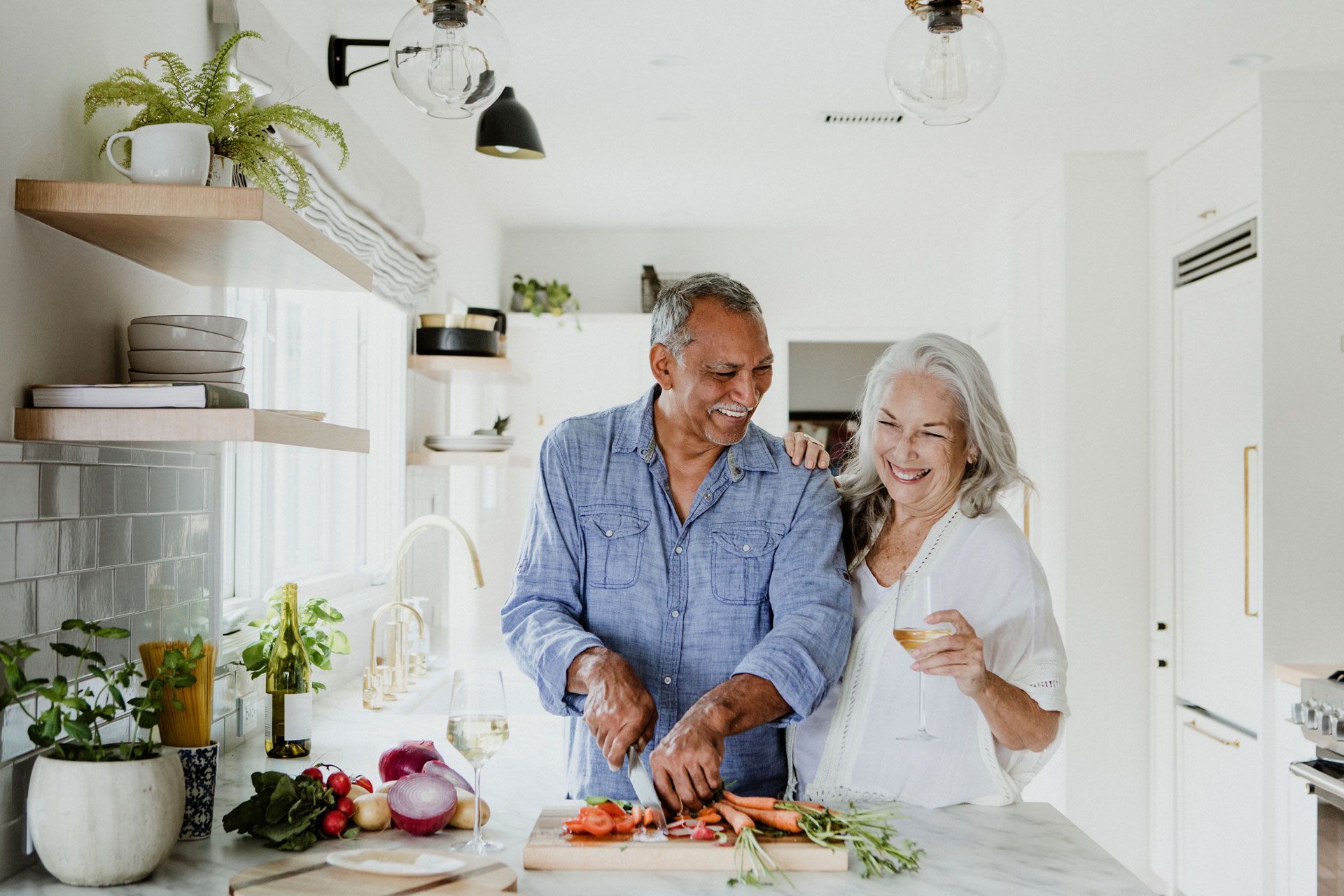 Couple smiling and preparing food together in a bright kitchen. Man chops vegetables; woman holds a piece, leaning in.
