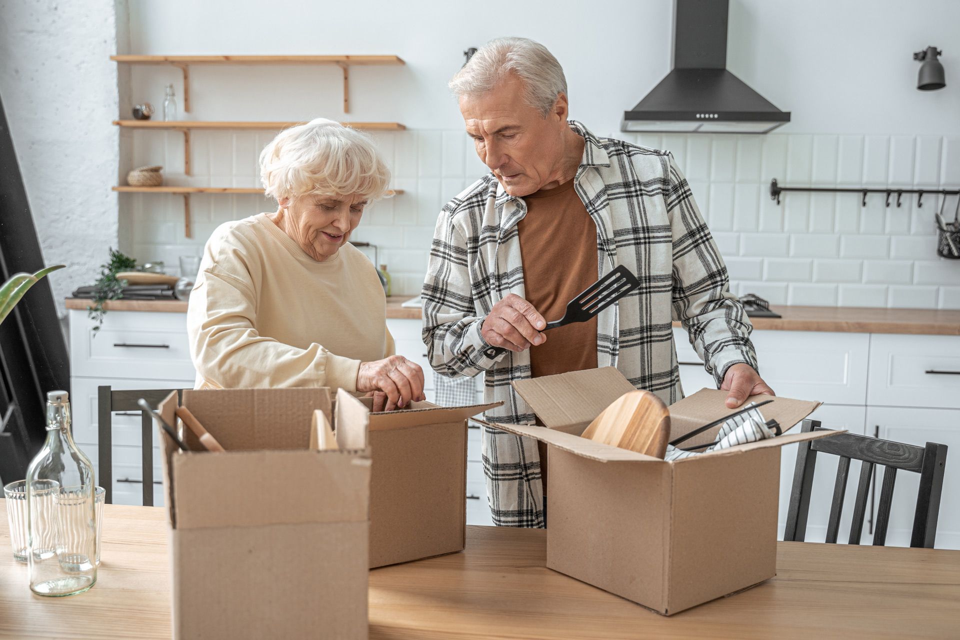 Older couple packing kitchen items into boxes in a well-lit kitchen.