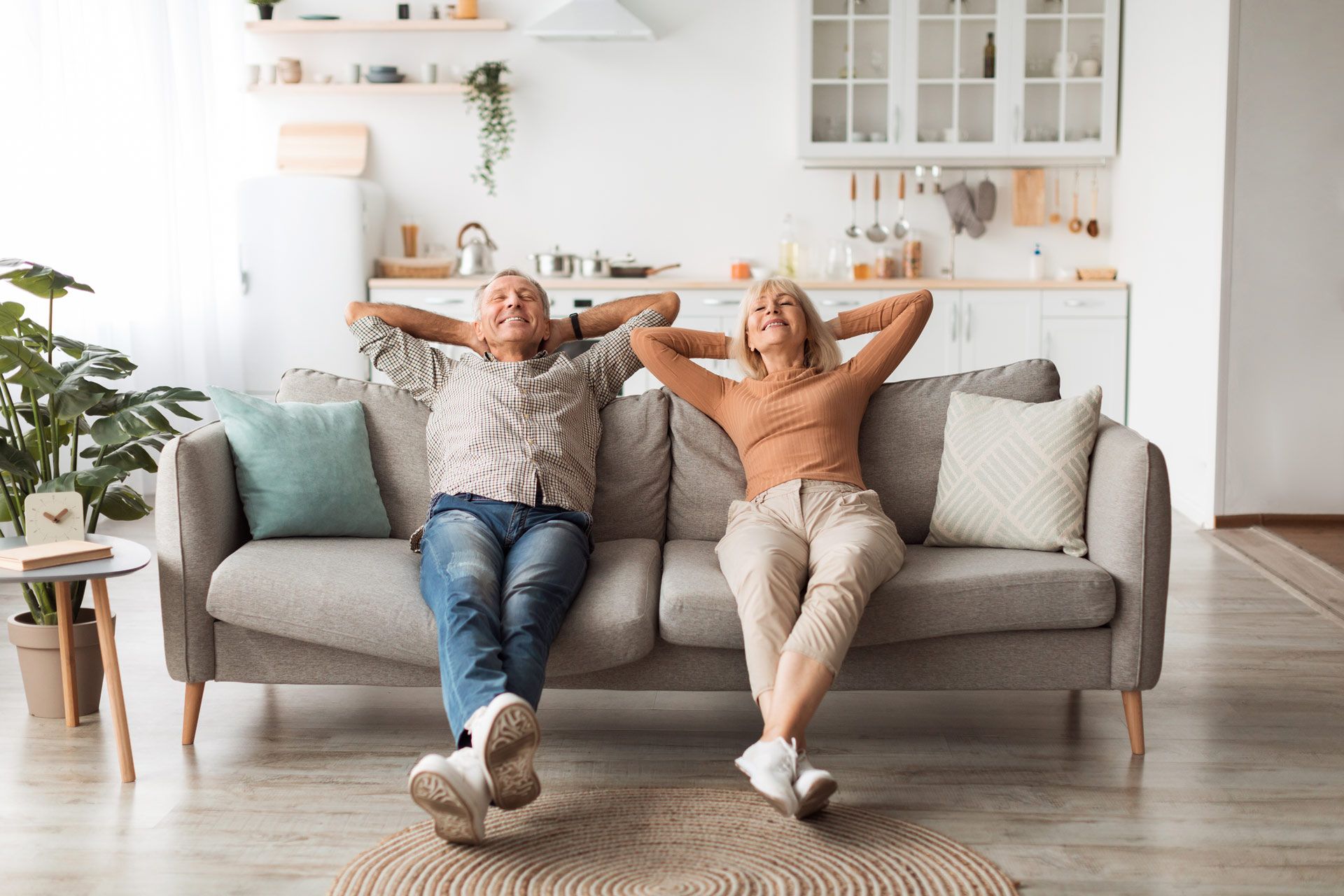 Couple relaxing on a couch with hands behind their heads, in a bright living room.