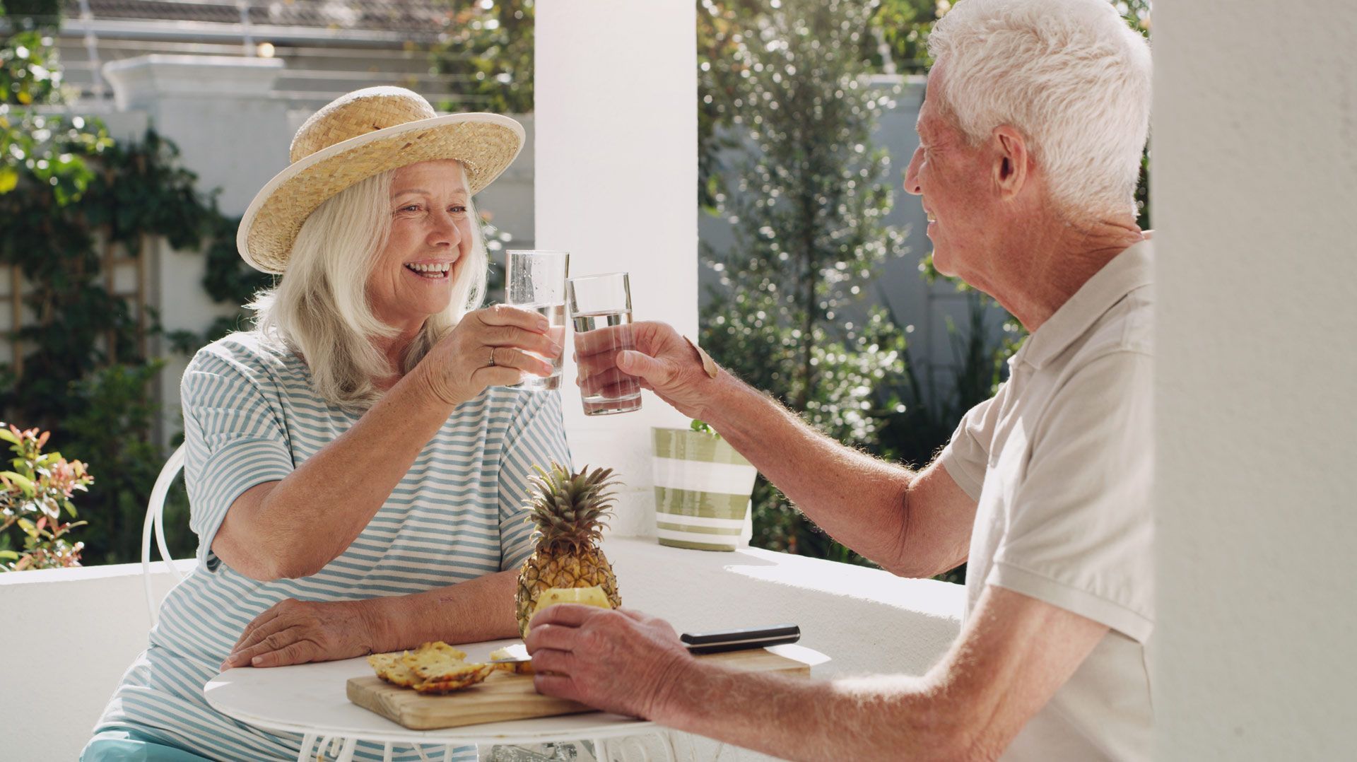 Elderly couple cheers with drinks on a sunny patio, smiling and enjoying a snack with pineapple.