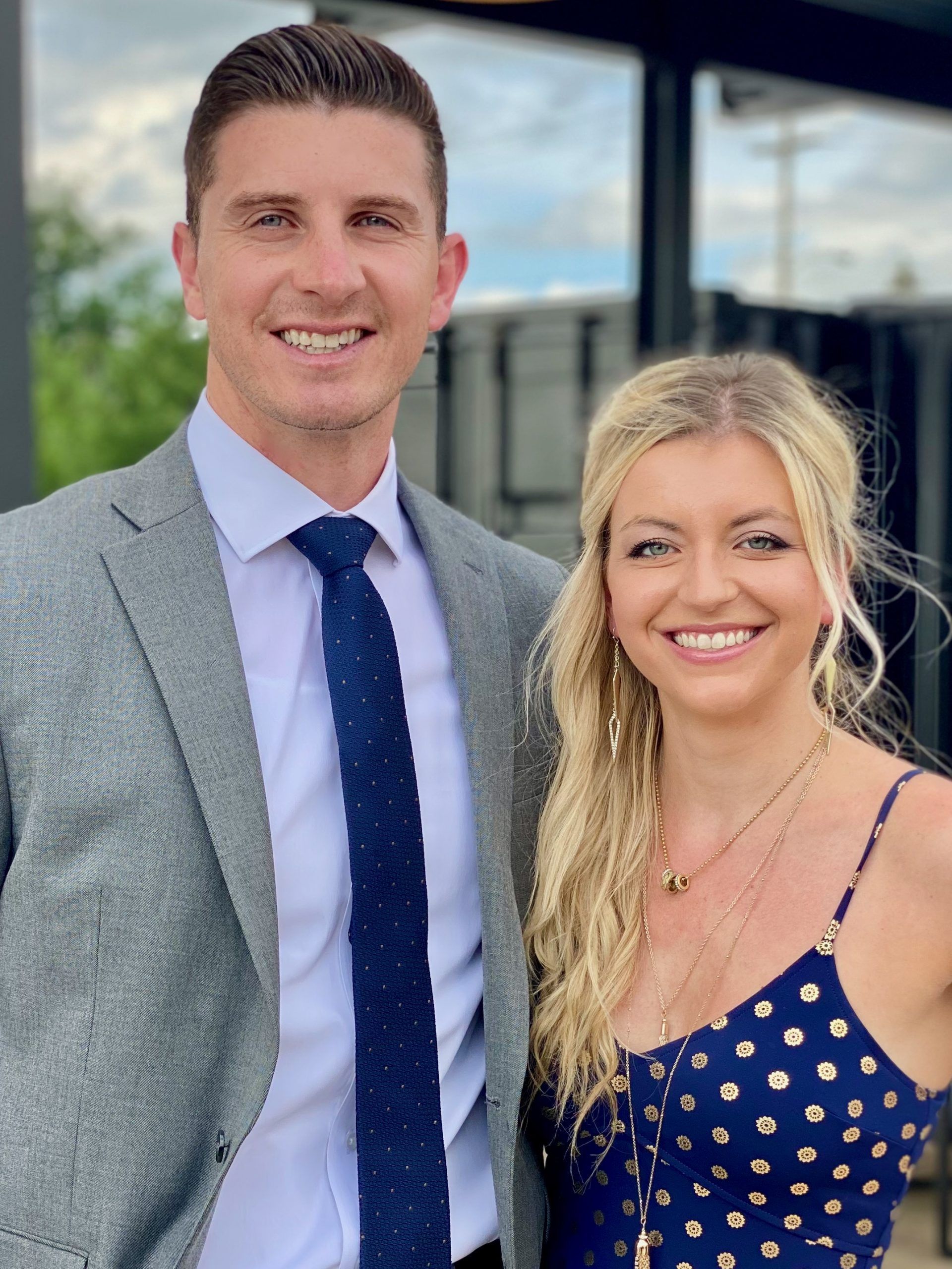 Man in grey blazer and woman in blue dress smile, standing outside.