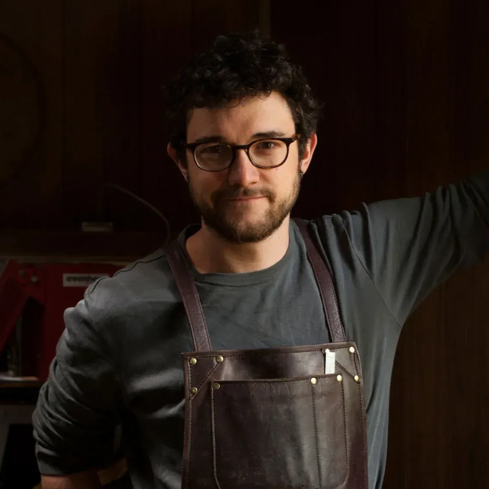 Man in apron, glasses, and grey shirt, leaning against a dark wood surface.