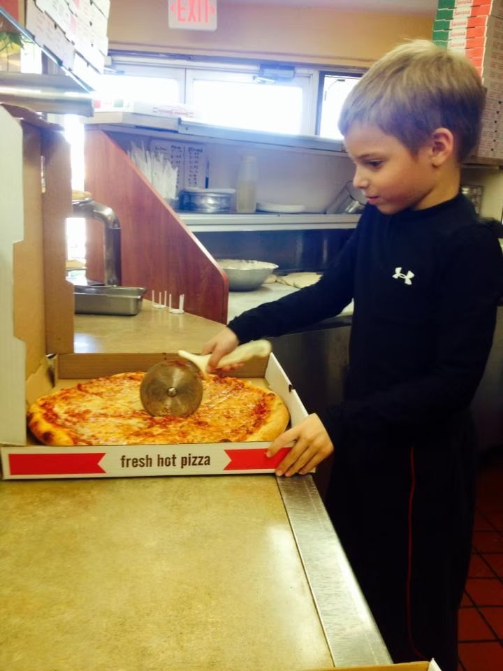 Boy cutting a fresh, hot cheese pizza with a wheel cutter in a pizza shop.