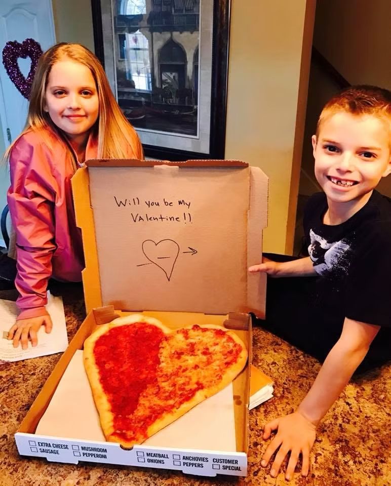 Two children holding a heart-shaped pizza in a box with a 