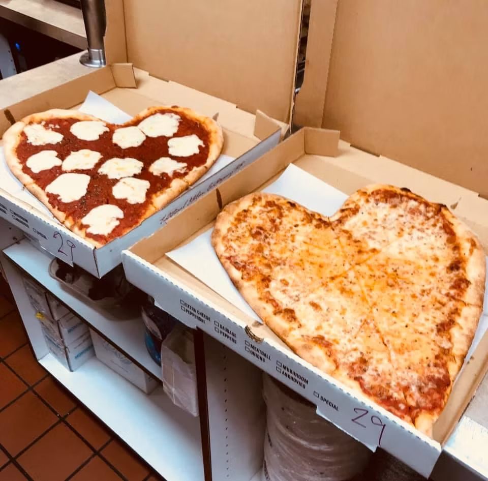Two heart-shaped pizzas in boxes: one with sauce and cheese, the other with cheese only, on a shelf.
