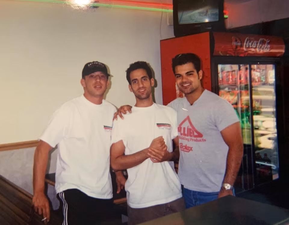 Three men in a restaurant pose for a photo. A Coca-Cola cooler is visible in the background.