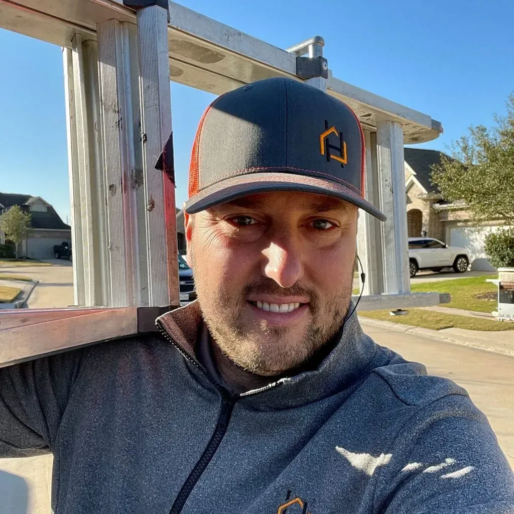 Man in cap with a logo smiles, carrying a ladder outdoors on a sunny day.