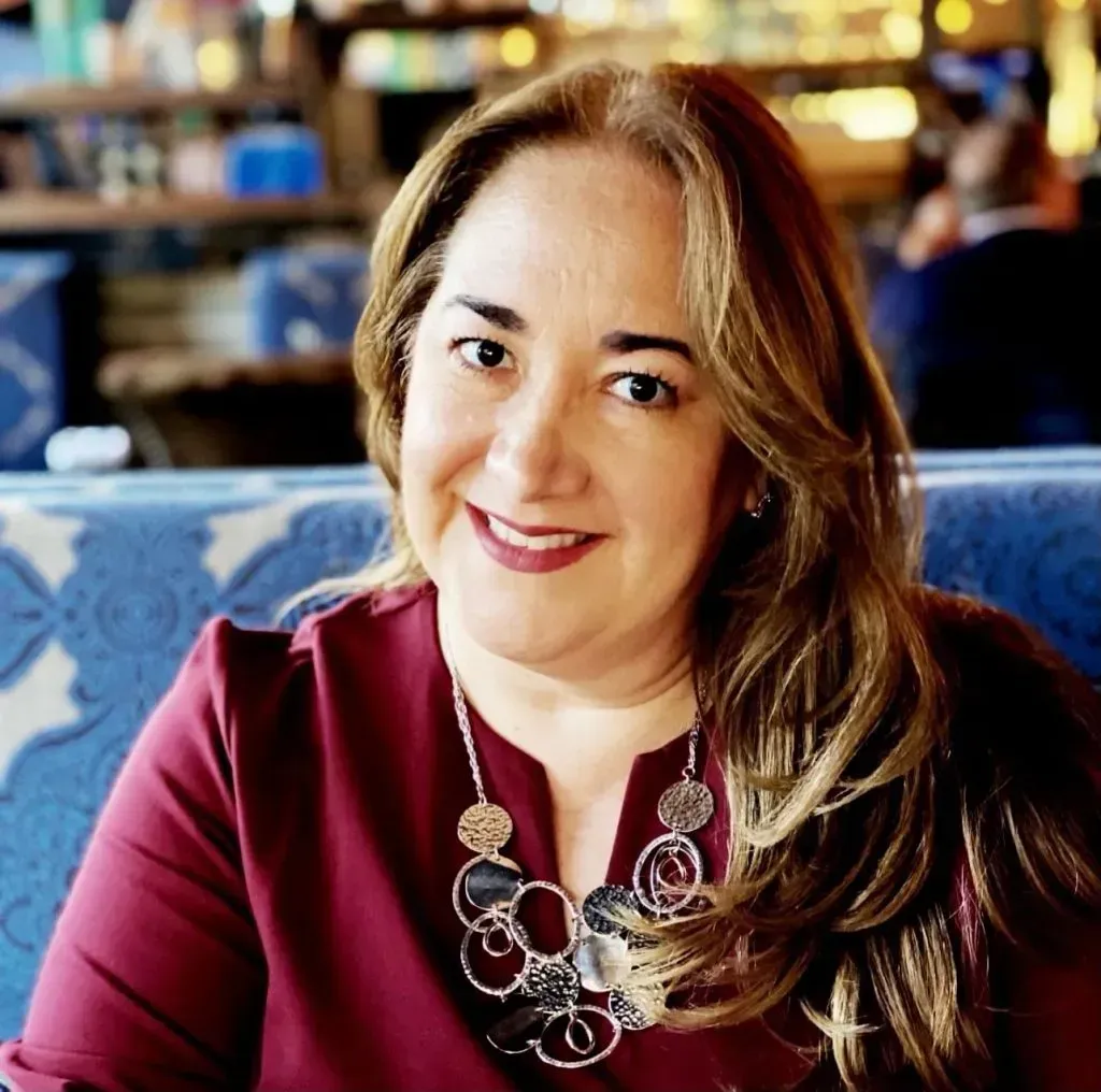Woman with long wavy hair smiles, wearing a burgundy top and silver necklace. Sitting at a table with a blue patterned back.