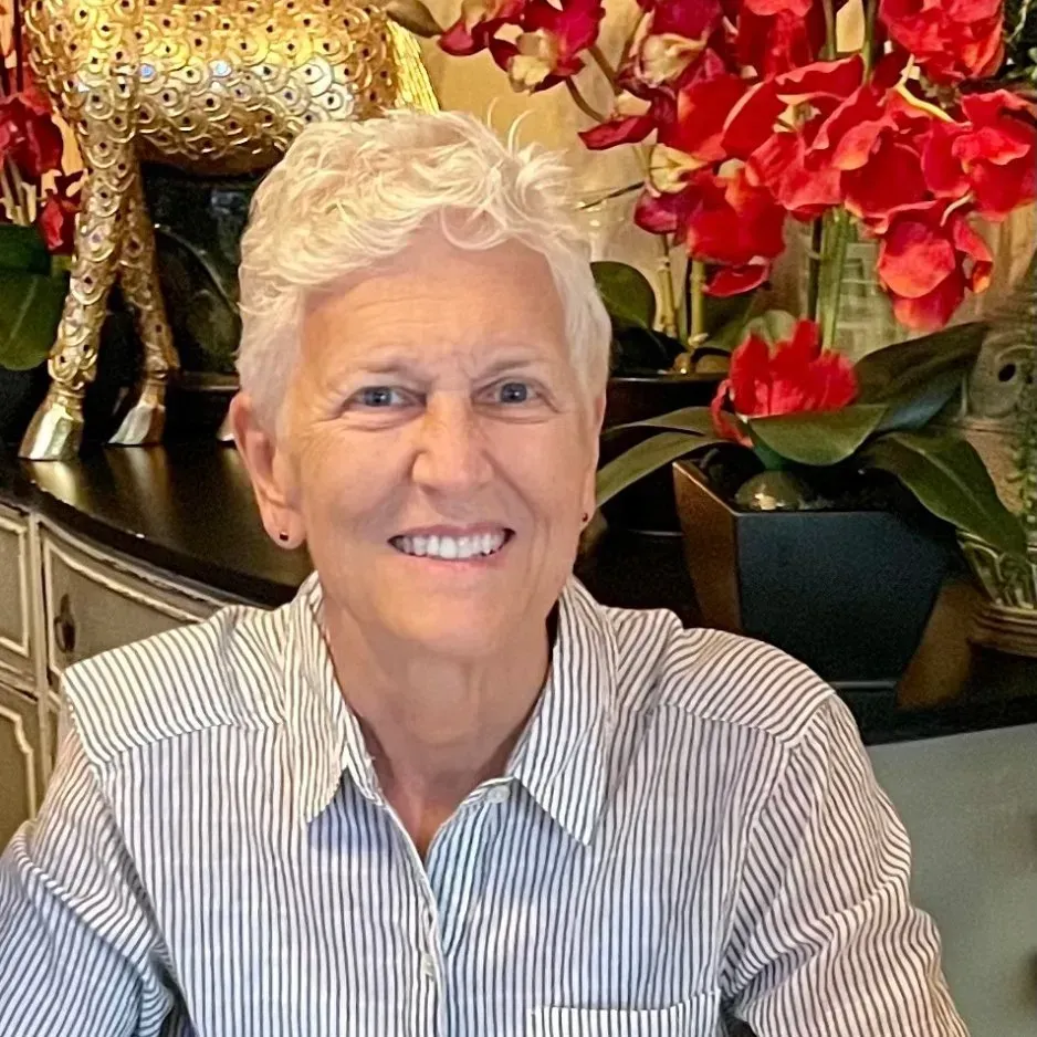 Woman with short white hair smiles, wearing a striped shirt. Red flowers in the background.