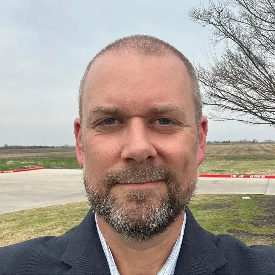 Man with a beard in a suit smiling at the camera, outdoors in front of a field and overcast sky.