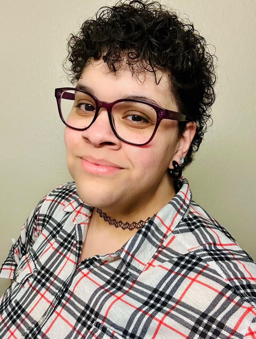 Person with short curly hair, glasses, and a plaid shirt, smiling at the camera.