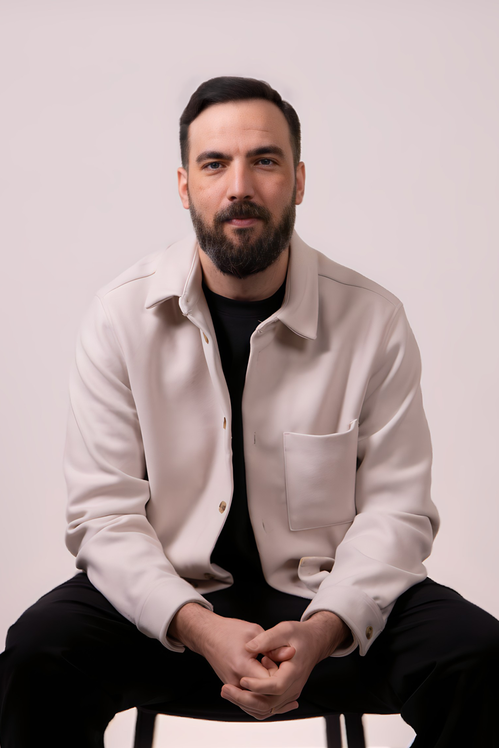 Man with a beard, seated, wearing a light jacket over a black shirt and dark pants; facing forward, neutral expression.