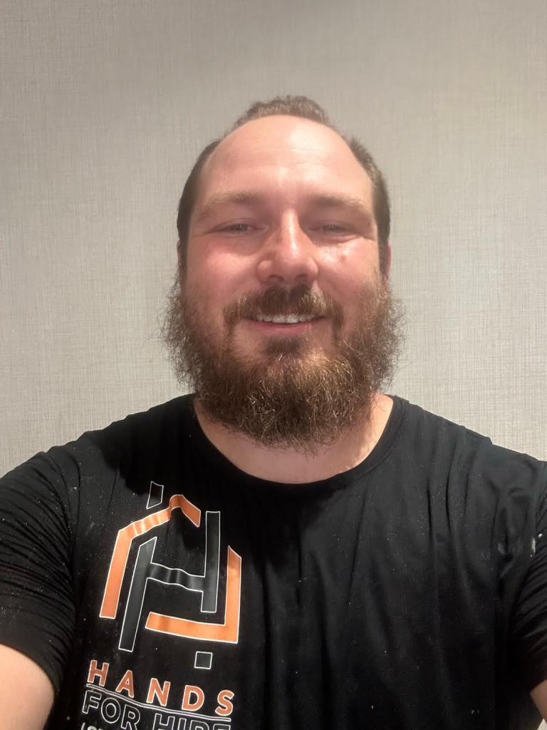 Man with beard wearing a black t-shirt with a logo, smiling.
