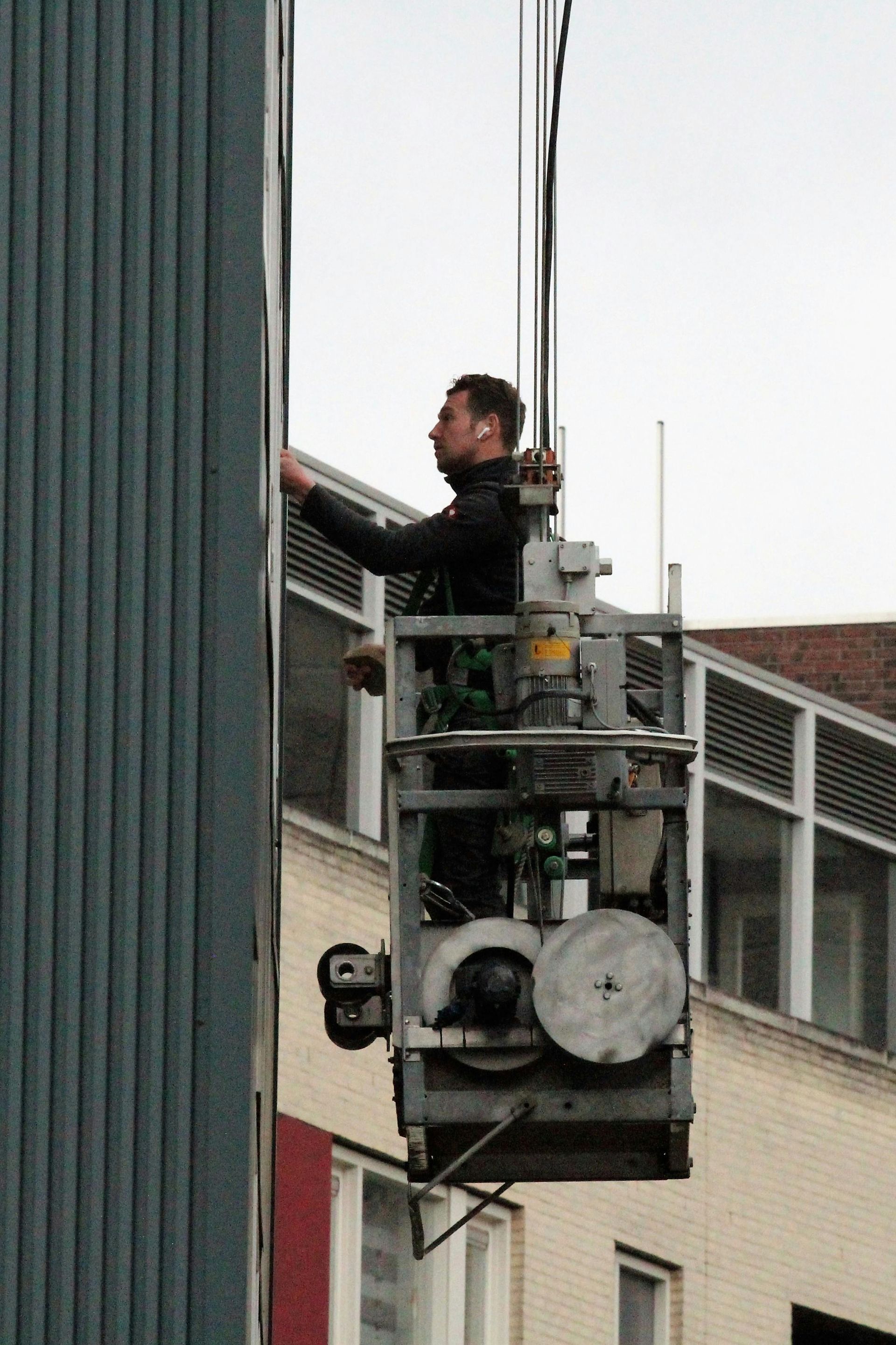 A worker in a suspended platform is inspecting a building facade. The platform is next to a blue wall.