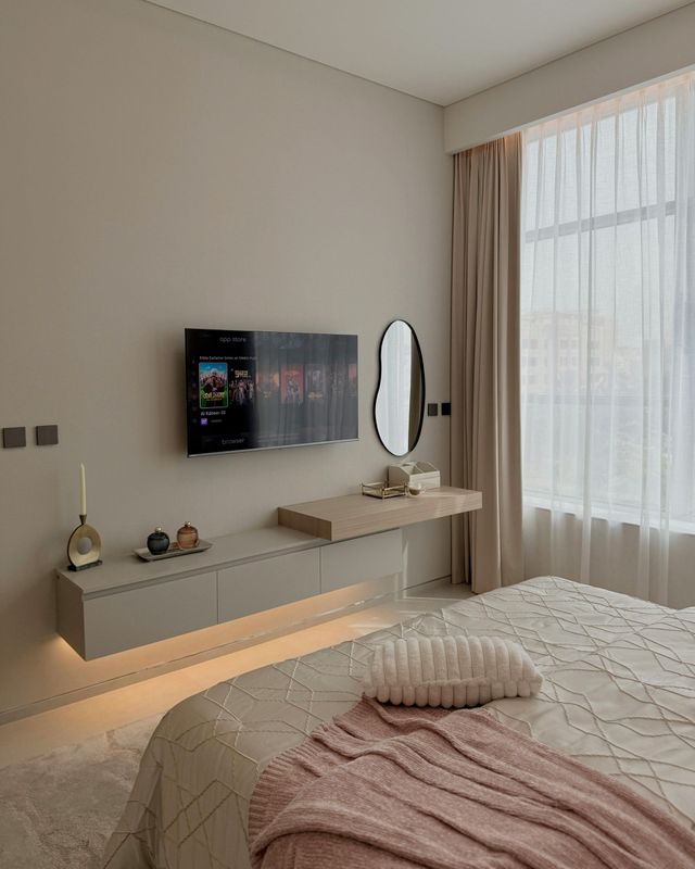 Modern bedroom with floating TV, vanity, and sheer curtains; beige and white color scheme.