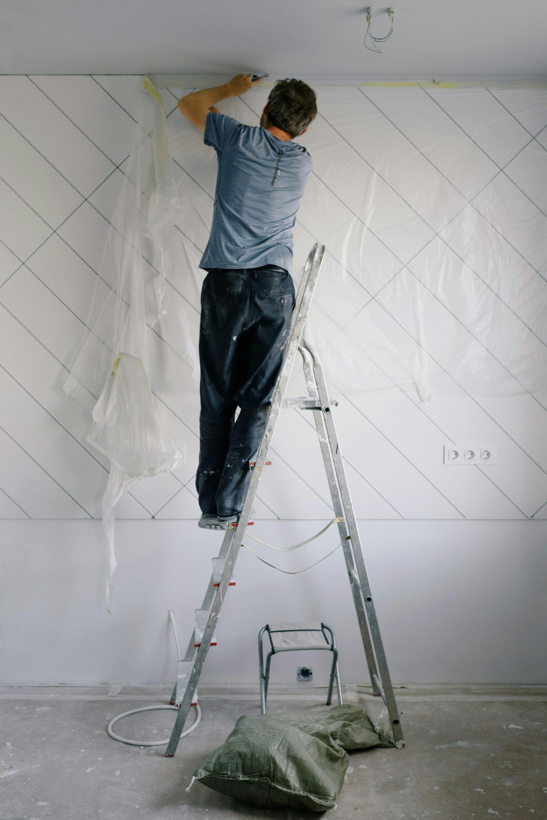 Person on a ladder tapes a wall with a geometric pattern. White wall, plastic sheeting, and green bag on the floor.