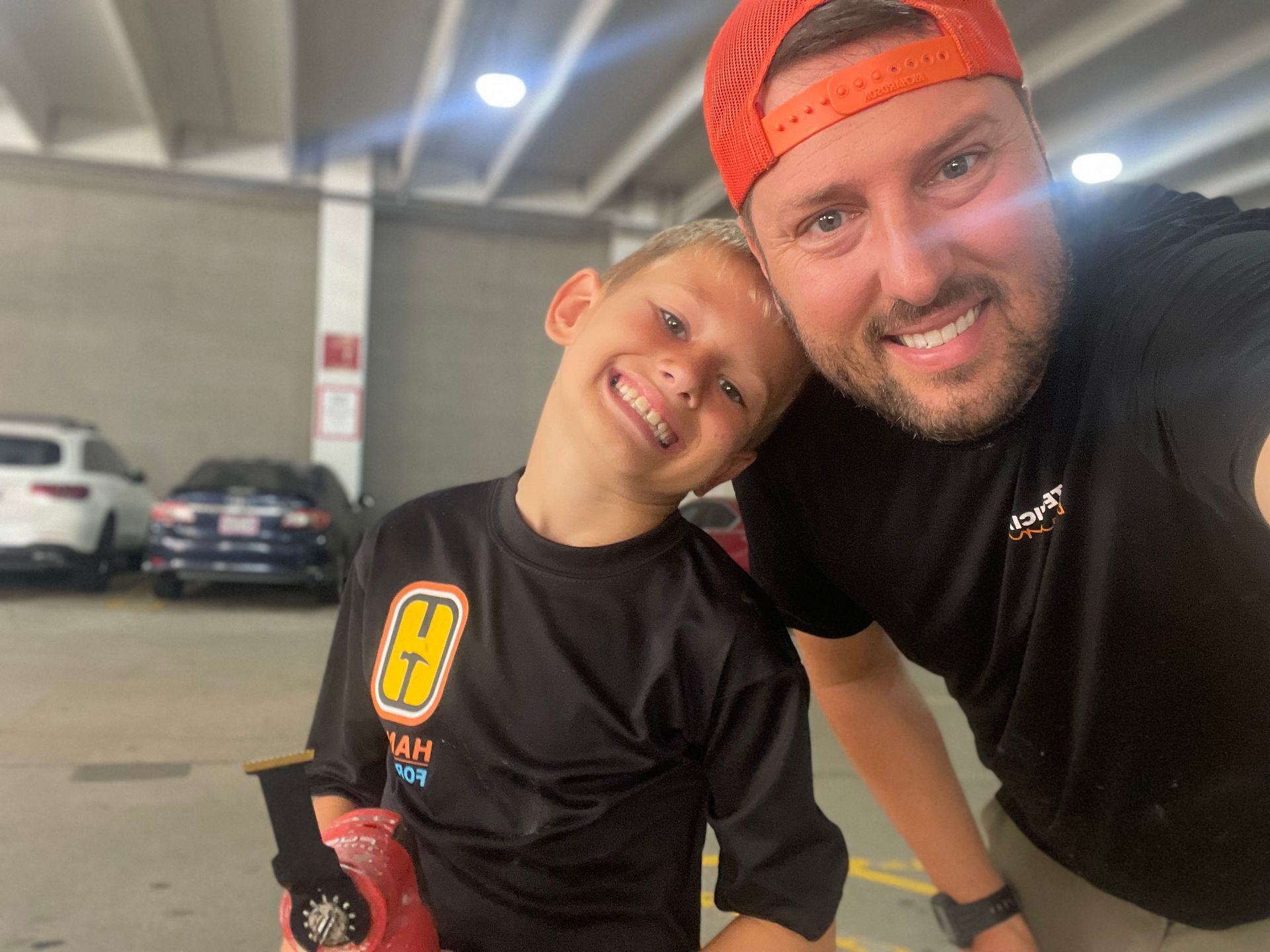 Man and boy smile for a selfie in parking garage. Boy in black jersey holds a toy. Man wears orange hat.