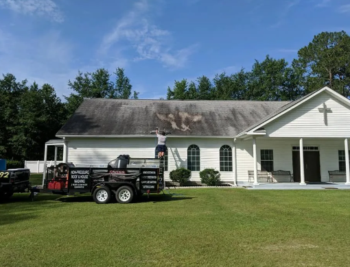 Man on ladder cleaning a church roof. Trailer and equipment on the lawn.