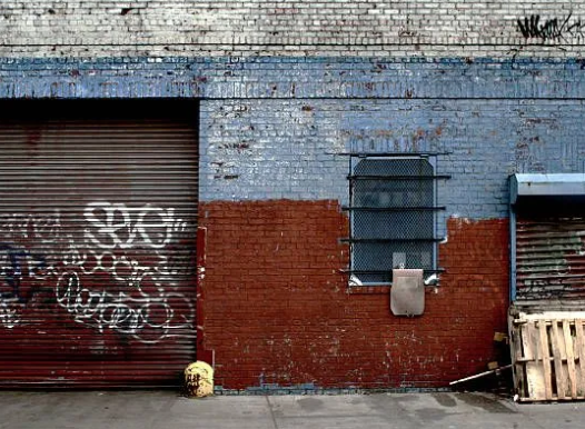 Brick building facade with graffiti, a grated window, and roll-up doors. Painted blue and red.