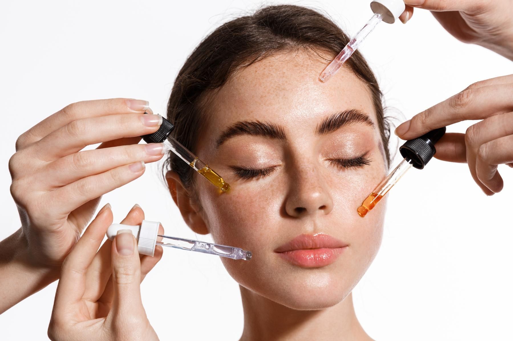 Woman with closed eyes receiving facial serum drops from multiple droppers against a white background