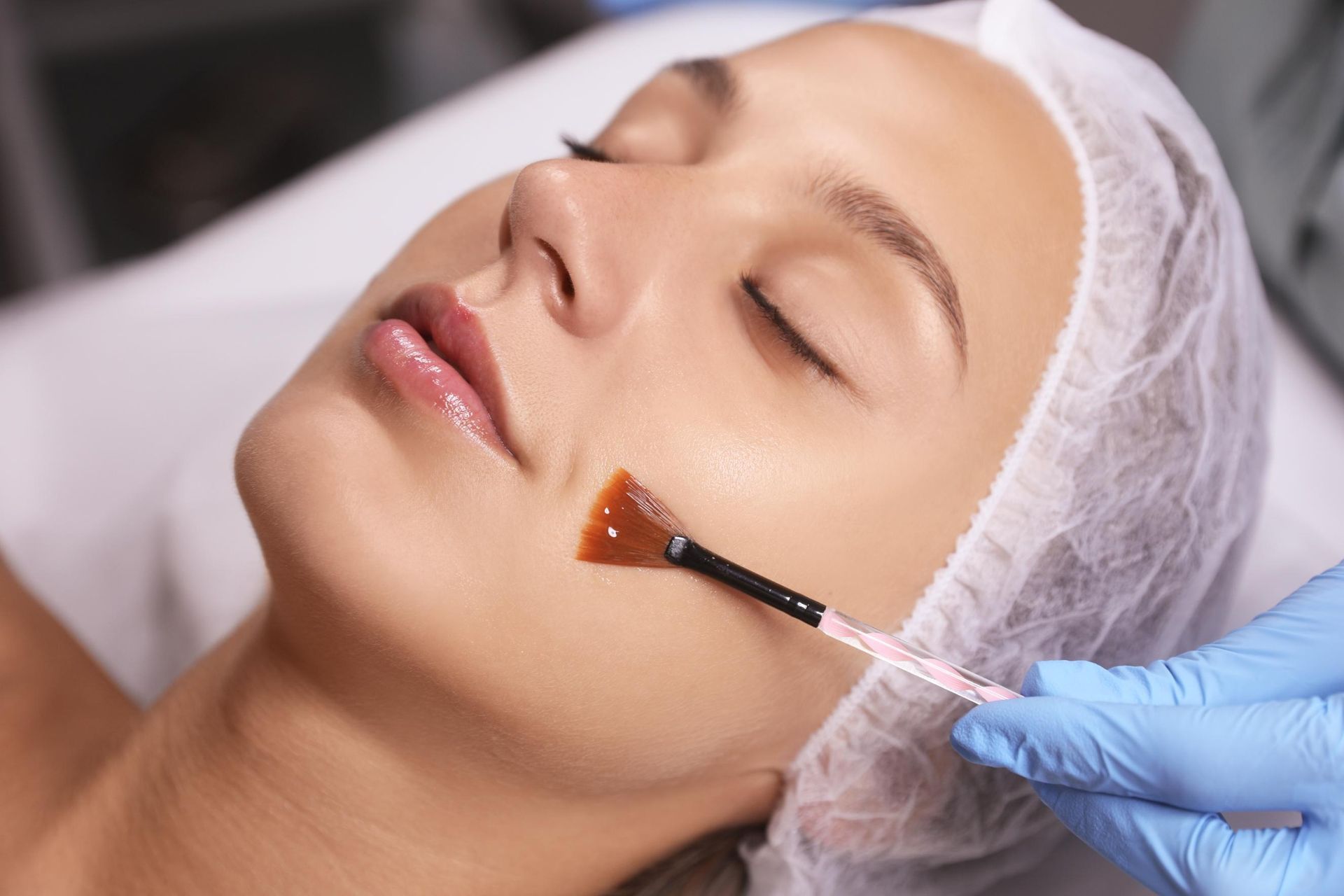 Gloved hand applying amber serum with a brush to a woman’s cheek in a spa treatment