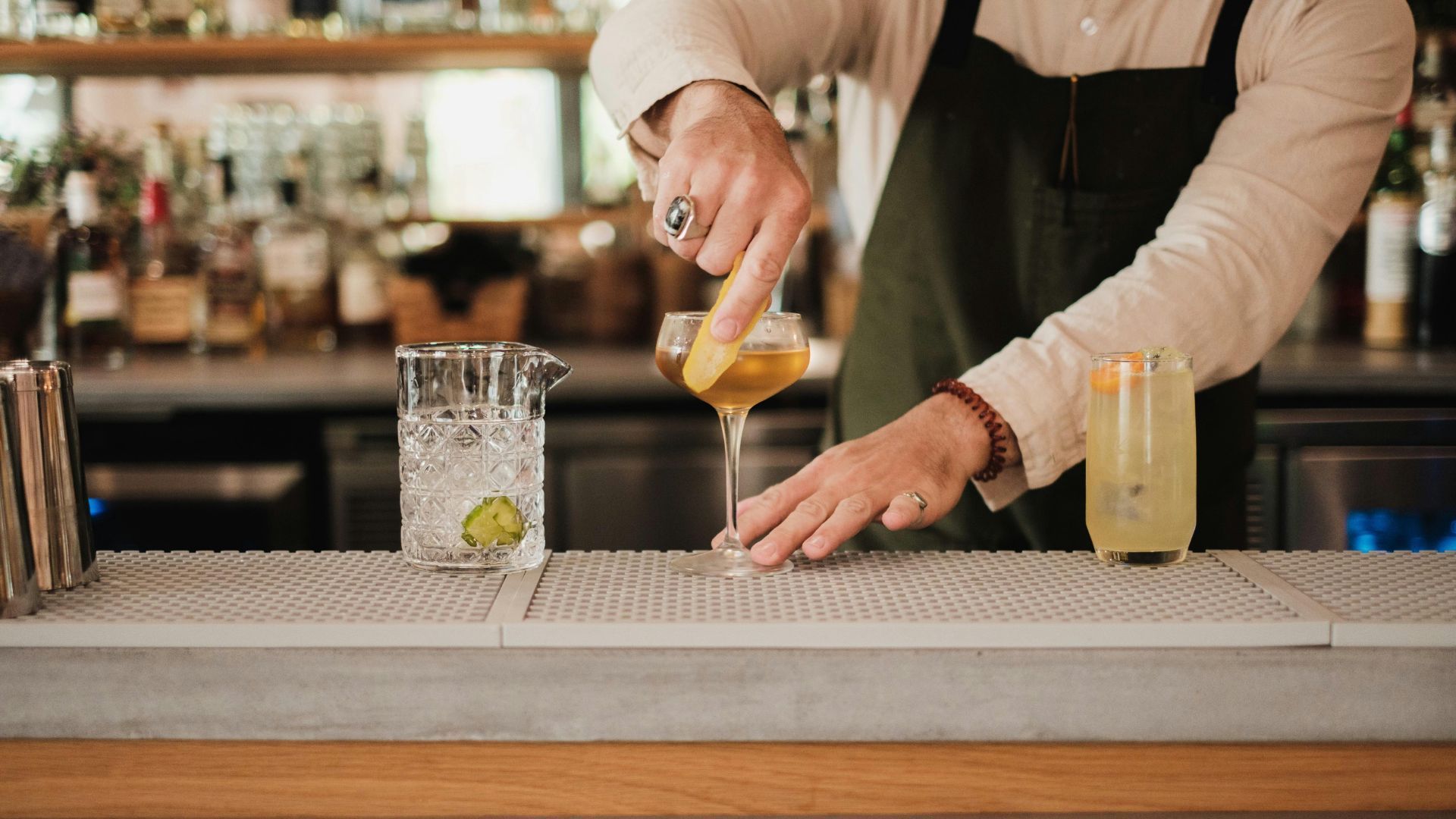 A bartender in an apron garnishing a cocktail with a citrus peel at a bar counter with bottles in the background.