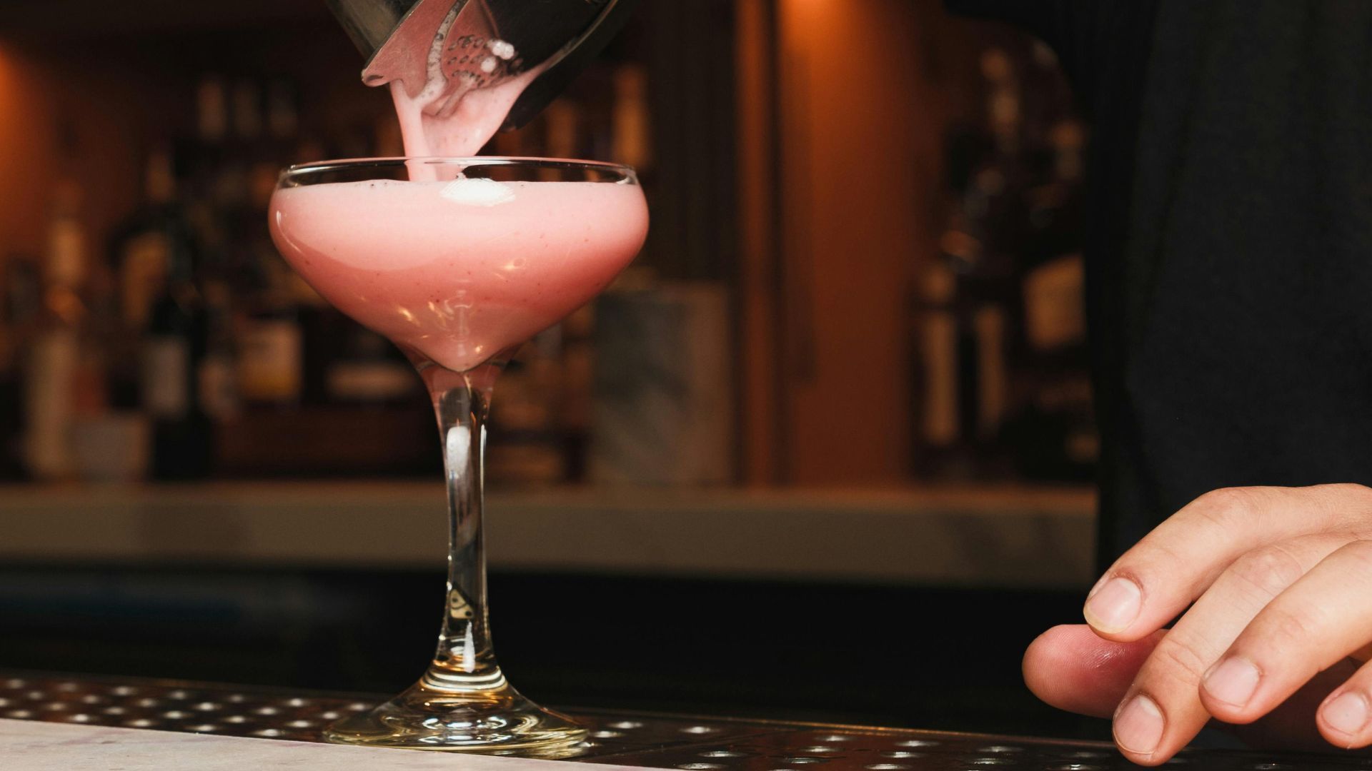 A bartender pours a frothy, pale pink cocktail into a stemmed coupe glass on a bar counter.