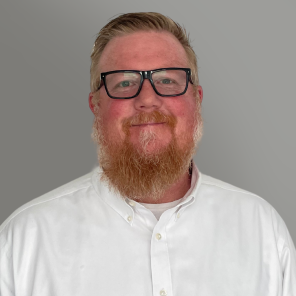 Smiling man with glasses and red beard in a white button-up shirt against a gray background