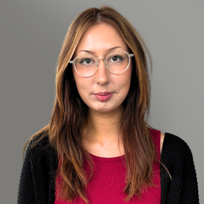 Portrait of a woman with glasses and long brown hair, wearing a magenta top and black cardigan against a gray background