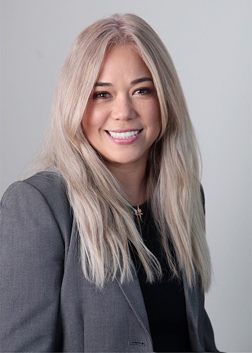 Smiling woman with long blonde hair in a gray blazer against a light background