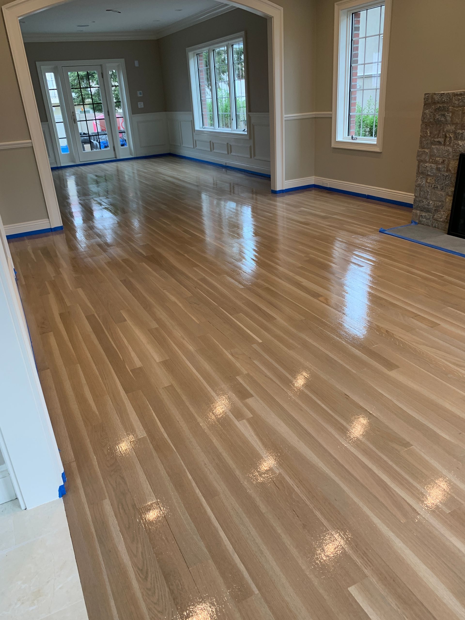 Newly refinished hardwood floors in a light-toned room with windows and blue painter's tape along the baseboards.