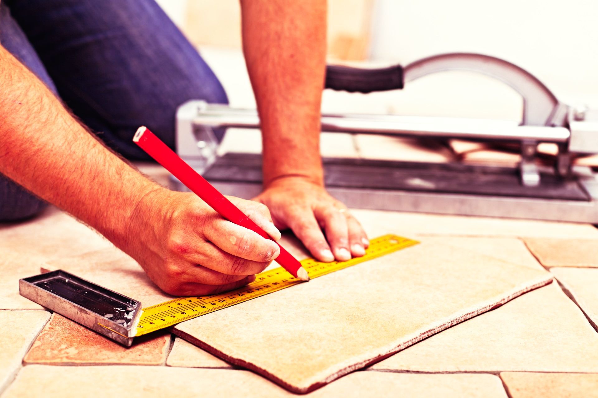 Person kneeling, measuring and marking a tile with a pencil and ruler; tile cutter in background.