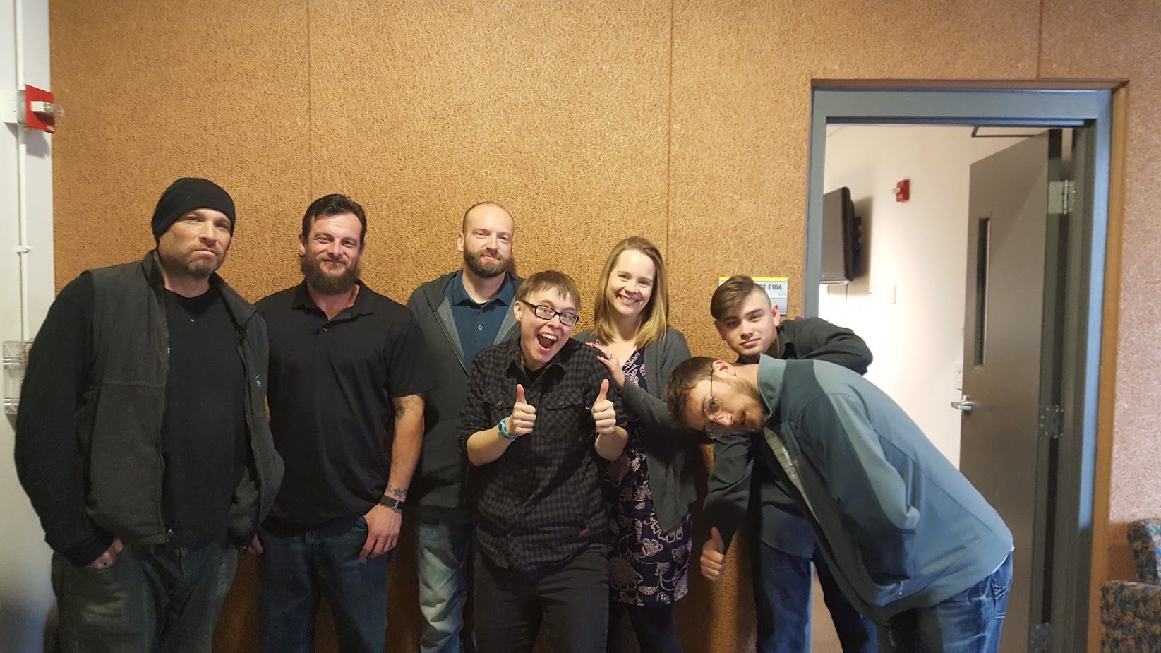 Five people posing together indoors against a tan wall, smiling and making playful gestures.